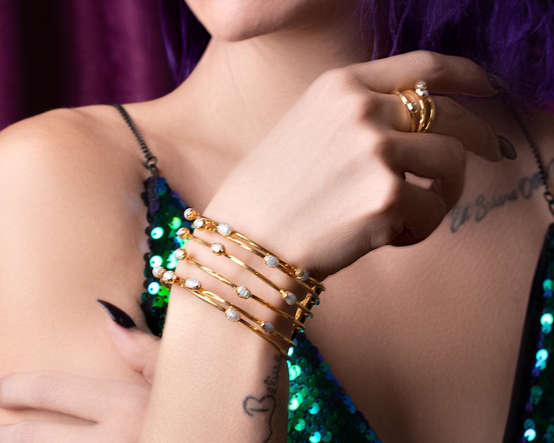 May include: A set of five gold bangles with white stones. The bangles are stacked on a person's wrist.