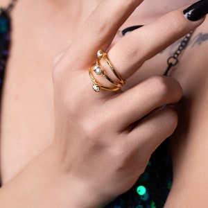 May include: A gold ring with three bands and pearl accents. The ring is worn on the index finger of a hand.