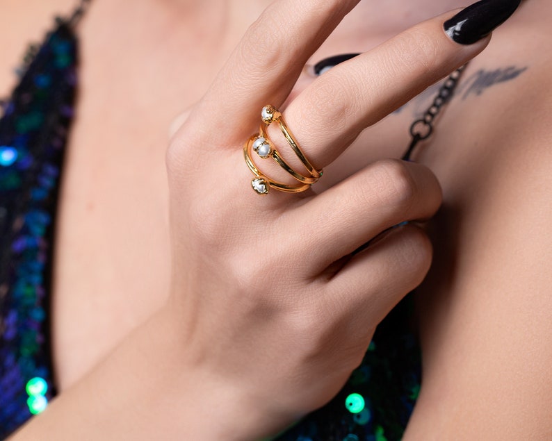 May include: A gold ring with three bands and pearl accents. The ring is worn on the index finger of a hand.