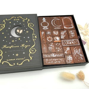 Könnte beinhalten: Ein schwarzes Box-Set mit der Aufschrift "Moonflower Magic" und einem Mond-Design. Im Inneren befinden sich zahlreiche Holzstempel mit himmlischen und botanischen Illustrationen. Das Set enthält Stempel von Monden, Sternen und floralen Designs.