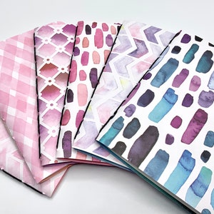 Könnte beinhalten: Sechs kleine Notizbücher mit verschiedenen Aquarellmustern. Die Cover zeigen rosa, lila und blaue Aquarelldesigns mit geometrischen Formen und abstrakten Mustern.