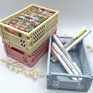 Könnte beinhalten: Zwei faltbare Plastikkisten in Beige und Rosa mit einem Stapel bunt gemustertem Papier in der oberen Kiste. Eine blaue faltbare Plastikkiste mit drei mehrfarbigen Textmarkern mit dem Text "Mildliner".