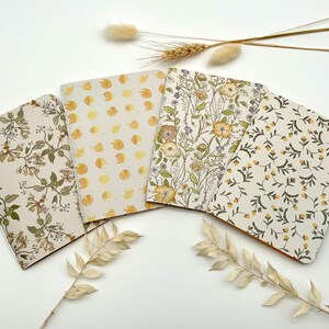 Könnte beinhalten: Vier Notizbücher mit floralen Mustern in Creme-, Gelb- und Grüntönen sind auf einer weißen Oberfläche angeordnet. Die Notizbücher weisen verschiedene botanische Designs auf, darunter Blumen und Blätter. Getrocknete Pflanzen sind um die Notizbücher herum platziert.