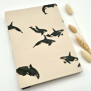 Könnte beinhalten: Ein beiges Notizbuch mit einem Muster aus schwarz-weißen Orcas. Die Orcas sind in verschiedenen Schwimmpositionen über dem Einband abgebildet. Das Notizbuch ist rechteckig und hat einen schwarzen Rücken.