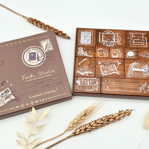 Könnte beinhalten: Ein braunes Set aus Holzstempeln mit weißen Illustrationen von Büchern, Fenstern und Schreibwaren. Die daneben liegende Box trägt den Text "Tsuki Studio" und "Notebook therapy". Dekorative Weizenähren befinden sich im Vordergrund.