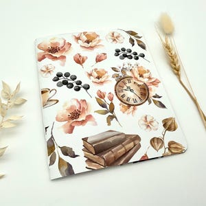 Könnte beinhalten: Ein rechteckiges Notizbuch mit weißem Einband und einem Aquarell-Druck von pfirsichfarbenen Blumen, dunkelbraunen Büchern, einer runden Uhr mit römischen Ziffern und dunklen Beeren. Das Design zeigt verschiedene botanische Elemente und Blätter.