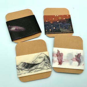 Könnte beinhalten: Vier dekorative Washi-Tape-Rollen mit einzigartigen Designs. Eine zeigt eine Galaxie, eine andere eine Sternennachtszene, eine dritte eine Bergketten-Skizze und die letzte abstrakte Figuren. Jede Rolle ist mit einer braunen Karte umwickelt.