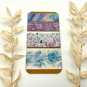 Könnte beinhalten: Ein Set aus vier dekorativen Washi-Tapes mit floralen und abstrakten Designs. Die Tapes weisen Aquarellmuster in Blau-, Lila- und Weißtönen auf. Die Tapes sind auf einer braunen Karte angeordnet, mit getrockneten Blättern auf beiden Seiten.