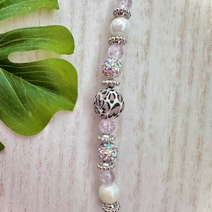 May include: A beaded charm featuring a string of decorative beads. The charm includes a large leopard print bead, iridescent beads, and smaller pink and silver beads. The charm is strung on a white cord.