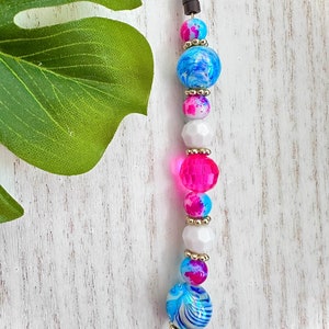 Puede incluir: Un collar de cuentas de colores con cuentas rosas, azules y blancas. Las cuentas son de vidrio y tienen un efecto marmoleado. El collar está enhebrado en un cordón marrón.