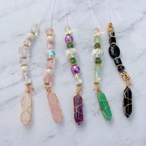 Puede incluir: Cinco colgantes de cristal con alambre dorado y cuentas. Cada colgante presenta un cristal de diferente color, incluyendo transparente, rosa, morado, verde y negro. Las cuentas son de varios colores y formas, incluyendo perlas y cristales iridiscentes.