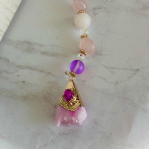 Puede incluir: Un amuleto de cristal rosa con un engaste chapado en oro y una piedra preciosa rosa. El amuleto está unido a una cadena de cuentas con cuentas rosas, moradas y transparentes.