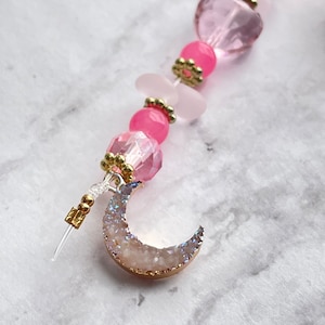 Puede incluir: Un amuleto de cuentas rosadas y doradas con un amuleto de media luna. El amuleto de la luna está hecho de cristal blanco con bordes dorados.