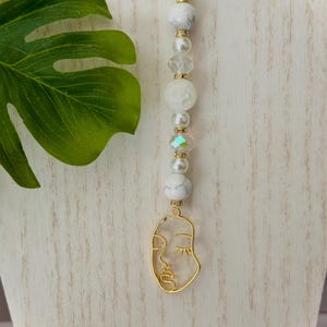 Pode incluir: Colar de contas brancas e douradas com um pingente de rosto dourado. O colar apresenta contas brancas, contas de cristal transparente e um pingente de rosto dourado.
