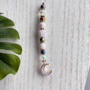 Puede incluir: Un dije de cuentas con un colgante de media luna. El dije presenta una cadena de cuentas iridiscentes, blancas y doradas. El colgante de media luna está incrustado con cristales brillantes. El dije está diseñado para colgarse.