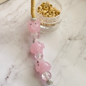 May include: A pink pig charm with clear and silver beads, strung on a thin cord. The charm features small, light-coloured cylindrical beads at the top. A small jar of light brown hair extensions is also visible.
