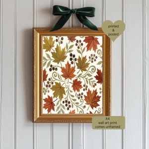 May include: An A4 wall art print featuring a pattern of orange, yellow, and brown maple leaves with small dark berries and light green sprigs on a cream background, displayed in a gold frame with a dark green ribbon bow at the top.