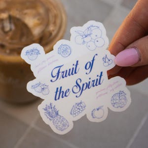 Könnte beinhalten: Ein weißer Aufkleber mit blauen Illustrationen von Früchten und dem Text "Fruit of the Spirit". Der Aufkleber wird von einer Hand mit rosa Nagellack gehalten, mit einem verschwommenen Hintergrund.