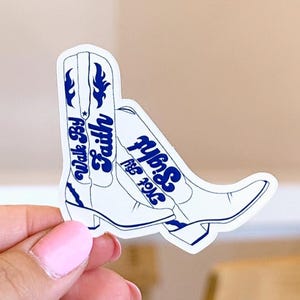 Könnte beinhalten: Ein weißer Aufkleber mit einem Paar Cowboy-Stiefeln mit blauen Akzenten. Die Stiefel tragen die Aufschriften "Walk By Faith" und "Not By Sight" in blauer Schrift. Der Aufkleber wird von einer Person gehalten.