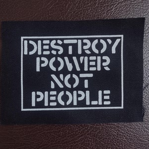 Peut inclure: Un écusson en tissu noir avec l'inscription blanche "DESTROY POWER NOT PEOPLE" en lettres de type pochoir. Le texte est encadré dans un rectangle blanc. L'écusson est posé sur une surface marron imitation cuir.