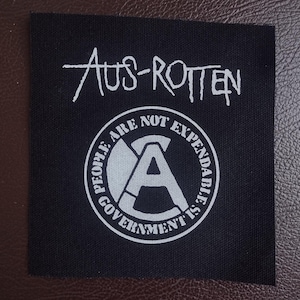 May include: Black and white patch with the text "Aus-Rotten" and a circle with the text "People are not expendable, government is" and a stylized letter "A" inside the circle.