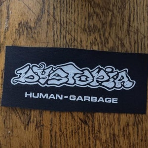 May include: A black rectangular patch with white text. The word "DYSTOPIA" is written in a stylized graffiti font above the phrase "HUMAN=GARBAGE". The patch is placed on a wooden surface.
