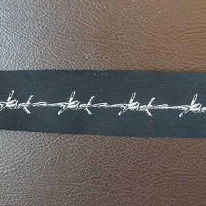 May include: A black fabric label with white barbed wire design. The label is rectangular and appears to be made of a woven material. The background is a textured brown surface.
