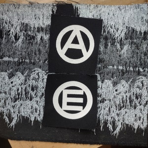 May include: Two black fabric patches with white embroidered symbols. The top patch has a circle with an 'A' inside, with a circle around it. The bottom patch has a circle with an 'E' inside, with a circle around it.