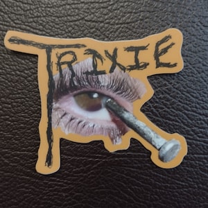 May include: A sticker with a yellow background and black text that says "TRIXIE". The sticker features a close-up of an eye with a nail through it.