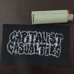 May include: Black and white embroidered patch with the text "CAPITALIST CASUALTIES" in a stylized font.