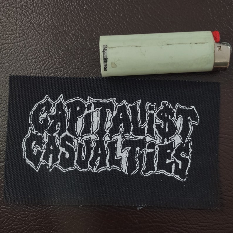 May include: Black and white embroidered patch with the text "CAPITALIST CASUALTIES" in a stylized font.