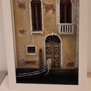 Puede incluir: Un modelo en miniatura enmarcado de un edificio con ventanas arqueadas y una puerta, sobre un fondo de canal con una góndola. El edificio está pintado en tonos amarillos y marrones, con molduras blancas. La góndola es negra y está parcialmente sumergida en el agua.