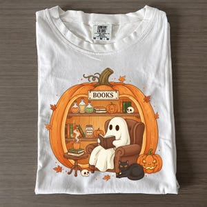 May include: A white t-shirt featuring a graphic of a large orange pumpkin carved into a bookshelf. Inside, a white ghost sits in a brown armchair reading a book, accompanied by a black cat, a steaming mug, and small pumpkins on shelves.