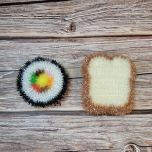 May include: Two crocheted dishcloths, one shaped like a sushi roll with colourful fillings and the other shaped like a slice of bread. Both dishcloths are white with brown edges.