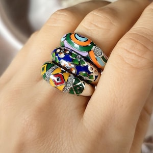 Puede incluir: Una mano muestra cuatro anillos de esmalte coloridos. Cada anillo presenta un diseño único con colores vibrantes como verde, naranja, azul y amarillo, acentuados con pequeñas piedras brillantes. Los anillos muestran patrones geométricos y florales.