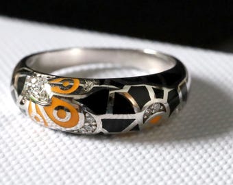 May include: Silver ring with a black enamel band, featuring orange and white decorative elements. The ring has a geometric design with small, clear gemstones. The ring is on a white surface.