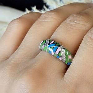 Op de afbeelding: Een kleurrijke ring met een geometrisch patroon in roze, groen, blauw en zwart. De ring is versierd met kleine, heldere edelstenen. Dit is een sieraad.