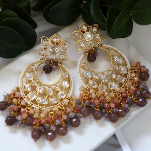 Pode incluir: Brincos de ouro com um design floral e formato de lua crescente. Os brincos são adornados com pedras transparentes e têm uma corrente pendente de contas rosa, roxas e marrons.