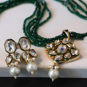 Pode incluir: Um colar de contas verdes com um pingente dourado de design quadrado com pedras transparentes e uma gota de pérola. Brincos combinando com pedras transparentes e gotas de pérola.