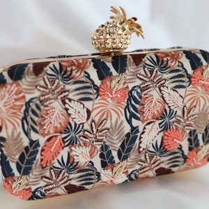 Op de afbeelding: Een beige clutch met een gouden ananasvormige sluiting. De clutch is versierd met een tropisch bladmotief in tinten blauw, groen en roze.