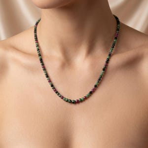 May include: A necklace of small, round beads in green, black, and red. The beads are strung together to form a necklace. The necklace is worn around the neck.