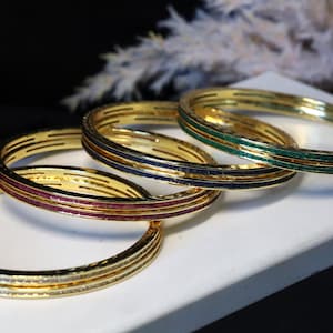 May include: Four gold bangles with different colored gemstones. The bangles are stacked on top of each other. The top bangle has green gemstones, the next has blue gemstones, the next has red gemstones, and the bottom bangle has clear gemstones.