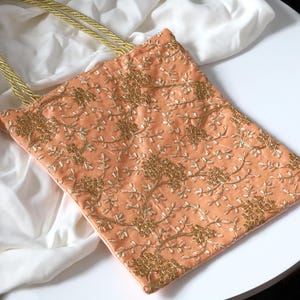 May include: Peach-coloured tote bag with gold floral embroidery and matching gold rope handles. The bag has a rectangular shape and is made of a soft, textured fabric. The floral design covers the entire surface of the bag.