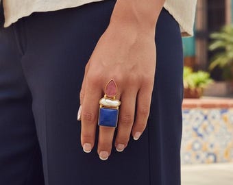 Könnte beinhalten: Ein goldfarbener Ring mit drei übereinander angeordneten Steinen: ein tropfenförmiger rosa Stein oben, ein ovaler weißer Stein in der Mitte und ein quadratischer blauer Stein unten. Der Ring wird am Finger getragen und zeigt die vertikale Anordnung.