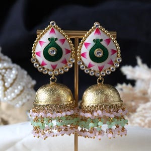 Pode incluir: Um par de brincos de ouro com um design floral branco e verde, acentos rosa e uma corrente pendente de contas verdes e rosa.