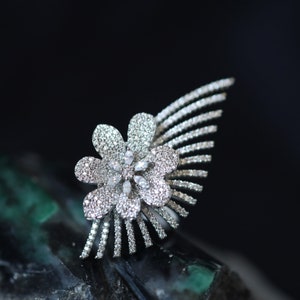 Puede incluir: Un anillo de plata con forma de flor con un diseño incrustado de diamantes. El anillo está colocado sobre un fondo verde oscuro.