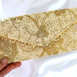May include: A cream-colored clutch with gold and white embroidery. The clutch has a pointed flap and a decorative trim of gold spikes.