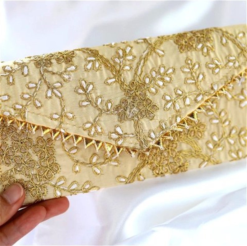 May include: A cream-colored clutch with gold and white embroidery. The clutch has a pointed flap and a decorative trim of gold spikes.