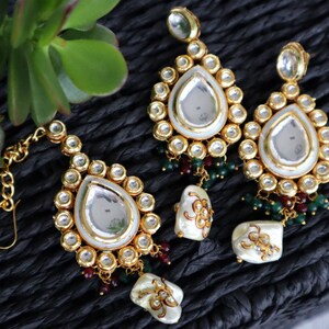 Pode incluir: Um conjunto de brincos e um piercing para o nariz dourados com detalhes em branco e verde. Os brincos têm um espelho em forma de lágrima rodeado por pequenas pedras redondas. O piercing para o nariz tem um design semelhante com um espelho e pedras menores.