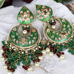 Pode incluir: Um par de brincos verdes e dourados com desenhos florais intrincados e contas penduradas. Os brincos têm a forma de uma meia lua com um acabamento de esmalte rosa e verde. As contas são uma mistura de verde, branco e vermelho.